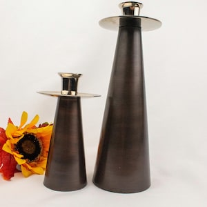 May include: Two bronze-colored candle holders with a conical shape. The candle holders have a gold-colored top and base. A sunflower and fall leaves are in the background. The candle holders are of different heights.
