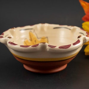 May include: A small, white ceramic bowl with a scalloped rim and a brown and yellow stripe design. The bowl has a stylized leaf design painted on the inside.