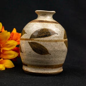 May include: A small, speckled ceramic vase with a round body and a narrow neck. The vase is off-white with brown leaf designs and horizontal bands. The background is black, with orange and yellow flowers on the left.