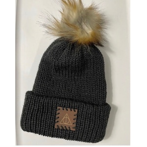 May include: Dark gray knit beanie with a faux fur pom-pom. A brown leather patch with a gold-colored symbol is sewn on the front. The hat is a winter accessory.