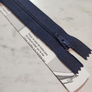 May include: A dark navy blue zipper with a matching pull tab, attached to a white paper instruction strip featuring black text and a small diagram. The text reads: Placer le pied-de-biche pour fermeture-éclair sur la machine. Faufiler à la machine sur.