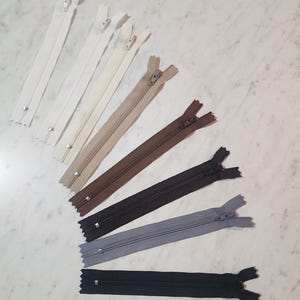 May include: A collection of seven nylon zippers arranged in a fan pattern, featuring a gradient of colors ranging from white and cream to beige, brown, dark brown, grey, and black, each with a metal pull tab and a small metal bottom stop.