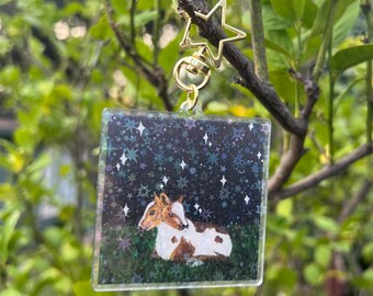 May include: A clear square keychain with a starry night background and a painting of two brown and white calves cuddling. The keychain has a gold star charm attached.