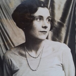 Puede incluir: Un retrato en blanco y negro de una mujer con un sombrero oscuro y un collar de perlas. Lleva una blusa blanca y tiene el pelo corto a la altura de los hombros.