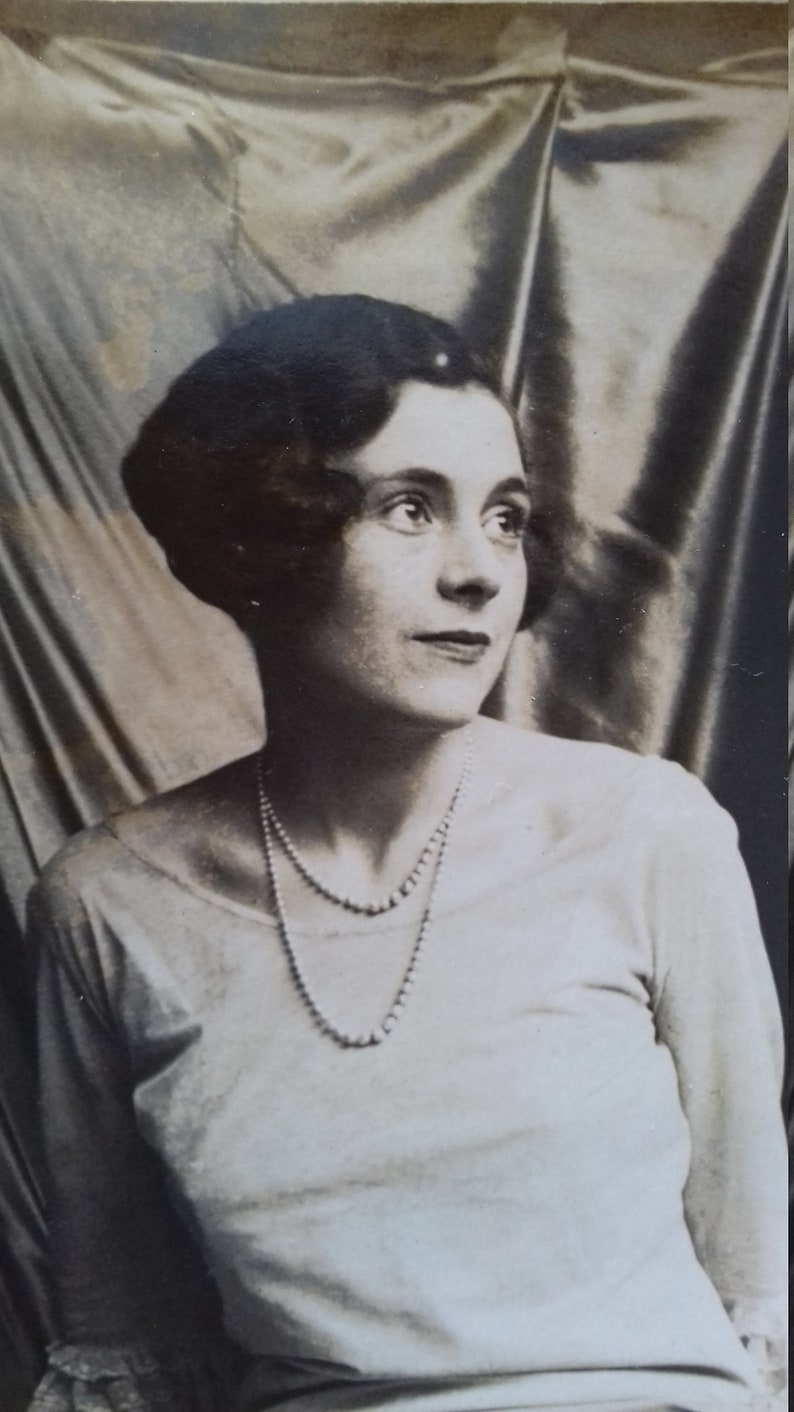 Puede incluir: Un retrato en blanco y negro de una mujer con un sombrero oscuro y un collar de perlas. Lleva una blusa blanca y tiene el pelo corto a la altura de los hombros.