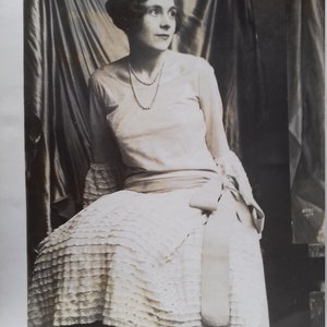 Puede incluir: Una fotograf&iacute;a en blanco y negro de una mujer con un vestido blanco con falda de volantes y un collar de perlas. Est&aacute; sentada en una silla con un fondo de tela drapeado.