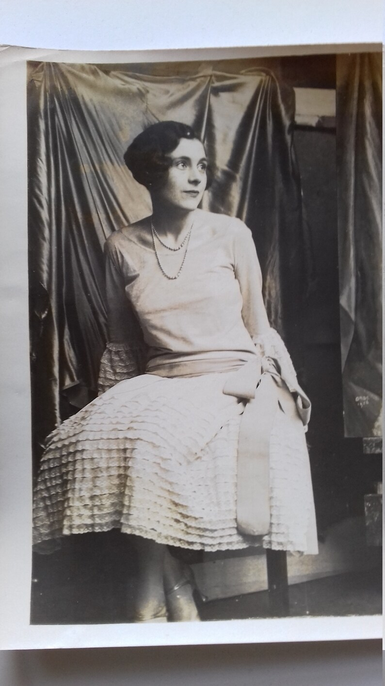 Puede incluir: Una fotograf&iacute;a en blanco y negro de una mujer con un vestido blanco con falda de volantes y un collar de perlas. Est&aacute; sentada en una silla con un fondo de tela drapeado.