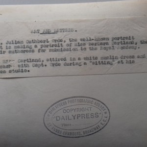 Puede incluir: Fotograf&iacute;a en blanco y negro de una mujer con un vestido blanco y una cinta azul. El texto en la parte posterior de la foto dice "Art and Letters. Capt. Julian Cuthbert Orde, el conocido retratista, est&aacute; haciendo un retrato de Miss Barbara Cartland, la autora de Mayfair, para su presentaci&oacute;n en la Royal Academy.  La Sra. Cartland, vestida con un vestido blanco de sol y una cinta azul con el Capt. Orde durante una "sesi&oacute;n" en su "estudio belsen."  Dailypress Photographic Agency. Copyright "Dailypress" St. Annes Chambers, Broadway, E.C.A."
