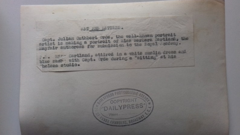 Puede incluir: Fotograf&iacute;a en blanco y negro de una mujer con un vestido blanco y una cinta azul. El texto en la parte posterior de la foto dice "Art and Letters. Capt. Julian Cuthbert Orde, el conocido retratista, est&aacute; haciendo un retrato de Miss Barbara Cartland, la autora de Mayfair, para su presentaci&oacute;n en la Royal Academy.  La Sra. Cartland, vestida con un vestido blanco de sol y una cinta azul con el Capt. Orde durante una "sesi&oacute;n" en su "estudio belsen."  Dailypress Photographic Agency. Copyright "Dailypress" St. Annes Chambers, Broadway, E.C.A."