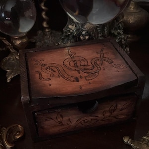 May include: A dark wooden box with a drawer, featuring an engraved design of a snake pierced by swords. The box has a rectangular shape and is stained in a warm brown color. The top of the box has a detailed carving.