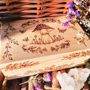 May include: A wooden box with a hand-burned design of a mushroom and foliage. The box is light brown and has a natural wood finish.
