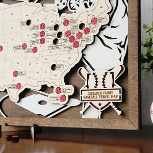 May include: A wooden map of the United States featuring laser-cut state borders and labeled baseball stadiums marked with red baseball icons. A decorative plaque reads "DECLERCO FAMILY BASEBALL TRAVEL MAP" over crossed bats and a baseball graphic.