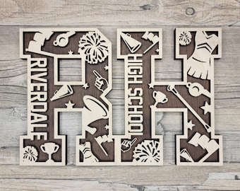 May include: A laser-cut wooden sign featuring the letters RH with the text Riverdale High School. The design includes cutouts of cheerleading pom-poms, megaphones, trophies, whistles, foam fingers, and flags against a dark brown background.