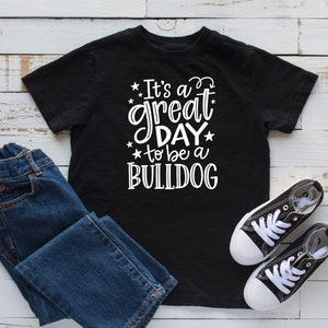 May include: Black t-shirt with white text that reads "It's a great day to be a BULLDOG". The shirt is laying on a white wooden surface with a pair of blue jeans and black sneakers.
