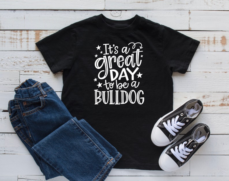 May include: Black t-shirt with white text that reads "It's a great day to be a BULLDOG". The shirt is laying on a white wooden surface with a pair of blue jeans and black sneakers.