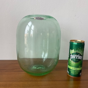 Può includere: Un vaso di vetro verde con una forma rotonda e bulbosa. Il vaso è appoggiato su una superficie di legno accanto a una lattina di Perrier Lime, acqua minerale gassata aromatizzata.