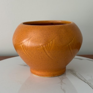 Pode incluir: Um vaso redondo de cerâmica laranja com uma textura salpicada e um padrão decorativo de folhas. O vaso tem uma abertura larga e um corpo arredondado, perfeito para exibir flores ou como peça decorativa.