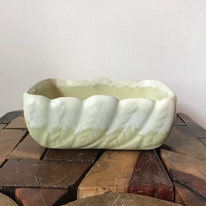 Pode incluir: Um vaso de cerâmica verde claro com uma borda ondulada e um esmalte branco. O vaso está sobre uma superfície de madeira.