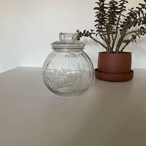 Puede incluir: Un tarro de cristal transparente con tapa, con forma de calabaza. El tarro tiene una cara de linterna tallada. El tarro está sobre una superficie blanca. Una planta en maceta está en el fondo.