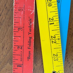May include: Three wooden folding rulers, one red, one yellow, and one blue. The red ruler has markings in inches and the text "Boye Folding Yardstick" printed on it. The yellow and blue rulers have markings in inches.