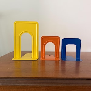 May include: Three metal bookends in descending sizes, featuring a smooth, glossy finish. The largest is bright yellow, the middle one is orange, and the smallest is blue. Each bookend has an arched cutout design and a flat base for stability.