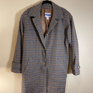 Op de afbeelding: Een vintage Pendleton-jas met pied-de-poule-motief in bruin-, blauw- en beigetinten. De jas heeft een klassieke kraag, een knoopsluiting en twee voorzakken. De jas hangt aan een zwarte hanger.