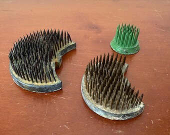 May include: Three floral frogs consisting of two crescent-shaped metal bases with numerous upright metal pins and one small circular green plastic base with a dense cluster of green plastic pins, all designed for arranging flowers in a container.