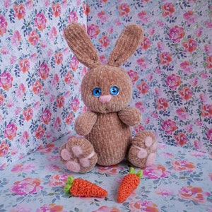 May include: A brown plush bunny rabbit with blue eyes and a pink nose sits on a floral patterned background with two orange carrots.