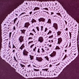 Puede incluir: Un tapete de ganchillo de color lavanda con un diseño geométrico calado. El tapete tiene un patrón circular central rodeado de bordes festoneados, creando una apariencia delicada y artesanal. El tapete es de forma octogonal.