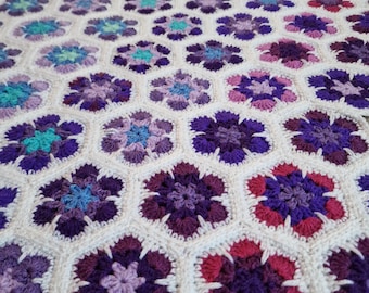 May include: A handcrafted crochet blanket with a floral design in shades of purple, pink, and teal. The flowers are arranged in a honeycomb pattern, with white yarn separating each hexagon. A cosy, decorative item.
