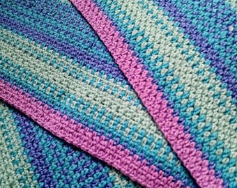 Puede incluir: Primer plano de una manta de ganchillo hecha a mano con un patrón de rayas. La manta presenta una paleta de colores de verde azulado, azul, morado y rosa. Los bordes están acabados con un borde rosa liso. La textura del hilo es visible.