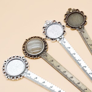 May include: Four metal bookmark rulers with decorative round frames. The rulers are in various shades of silver and bronze, with a scalloped edge and etched measurement markings. The frames have floral and ornate designs, with a blank circular space in the center.