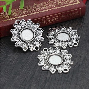 May include: Three silver-toned metal flower-shaped pendant charms with a round, open center for adding a cabochon. Each charm has a detailed design with a textured surface.