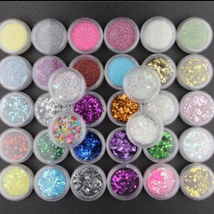 May include: A set of 24 small jars filled with various colours of glitter and sequins. The jars are arranged in a grid pattern on a black surface.