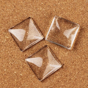 May include: Three clear glass square cabochons, also known as glass domes, on a cork background.