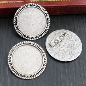 May include: Three silver-toned metal round blank cabochon settings with a decorative border. The settings have a pin back for attaching to clothing or fabric.