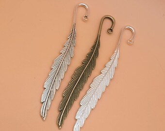 May include: Three silver feather-shaped bookmarks with a curled top. The bookmarks are in different shades of silver, with one being a darker bronze color.