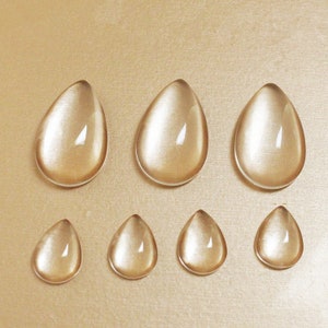 Könnte beinhalten: Sieben tropfenförmige, klare, bernsteinfarbene Glascabochons in verschiedenen Größen sind auf einer goldfarbenen Oberfläche angeordnet. Die Cabochons haben ein glattes, abgerundetes Aussehen mit subtiler interner Reflexion.