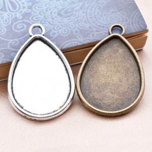 Könnte beinhalten: Zwei tropfenförmige Anhänger-Tabletts, eines silberfarben und eines bronzefarben, mit einer glatten, abgerundeten Kante und einer Öse oben zum Aufhängen.