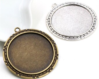 Op de afbeelding: Twee ronde hangerbakjes, één in antiek brons en de andere in zilver. Beide hebben een cirkelvormige verzonken ruimte voor het plaatsen van een ontwerp, met een decoratieve rand en een lus voor het bevestigen van een ketting of koord. Ideaal voor het maken van sieraden.
