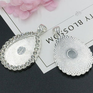 Könnte beinhalten: Zwei silberne Anhänger in Tropfenform mit einem dekorativen Design. Ein Anhänger ist mit klaren Strasssteinen am Rand verziert.