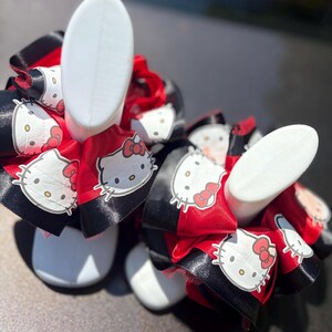 Pode incluir: Dois pares de formas de sapatos brancos são adornados com grandes laços. Os laços apresentam um esquema de cores vermelho e preto, com um padrão repetido do personagem Hello Kitty. Os laços são feitos de fita de cetim.