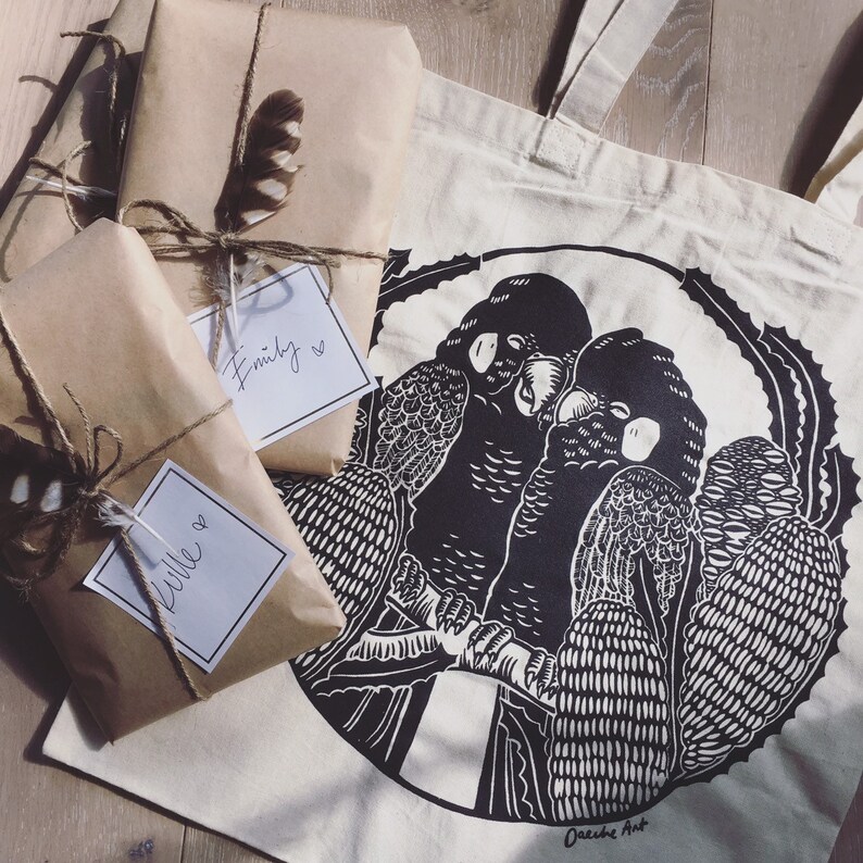 May include: A white tote bag with black handles features a black and white illustration of two birds perched on a branch. The bag has the text "Oasys Art" printed on the bottom. Two brown gift-wrapped packages with handwritten tags are in the foreground.