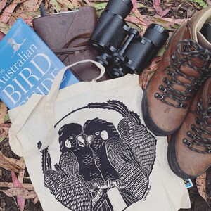 May include: A white canvas tote bag with black and white illustrations of two birds perched on a branch. The bag has the text "Cheers to the Art" printed on the bottom right corner.