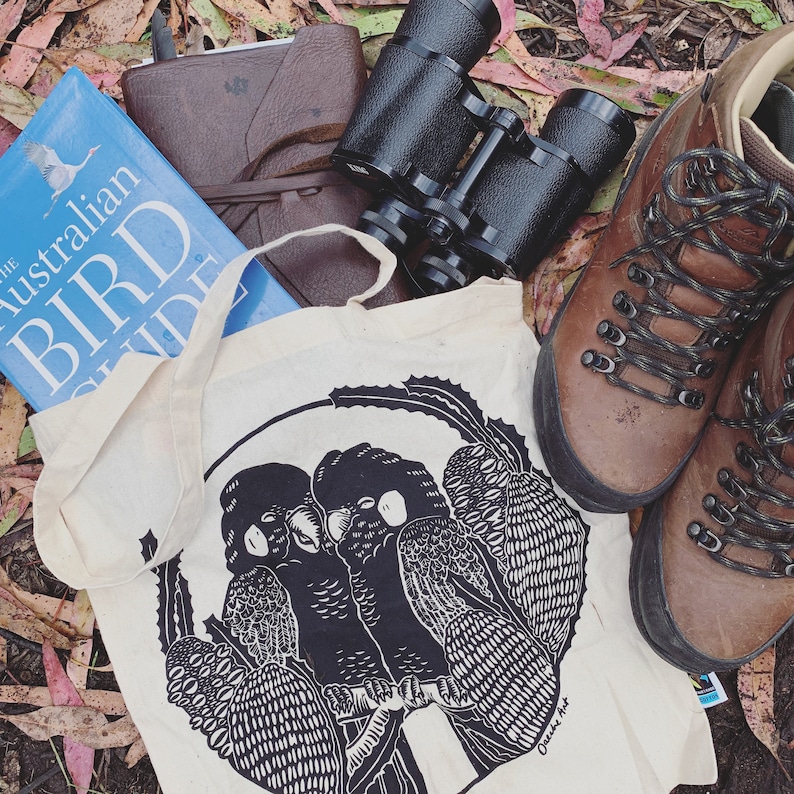 May include: A white canvas tote bag with black and white illustrations of two birds perched on a branch. The bag has the text "Cheers to the Art" printed on the bottom right corner.