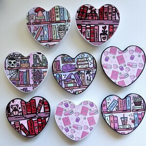 May include: Nine heart-shaped decorative items with various designs. Each features bookshelves with books, a cat, and other illustrations. The color scheme includes pink, purple, and white, with black outlines. The items are ideal for book lovers.