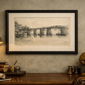 May include: A framed black and white print of a bridge over water hangs above a wooden desk. A brass desk lamp, books, and a globe are also on the desk. The print is in a black frame with a white mat.