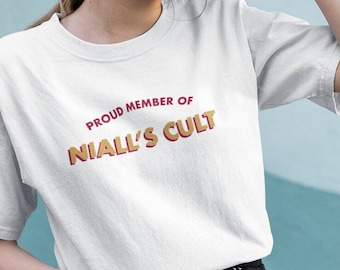 May include: White t-shirt with the text "PROUD MEMBER OF NIALL'S CULT" in red and gold lettering. The shirt is a classic crew neck style.
