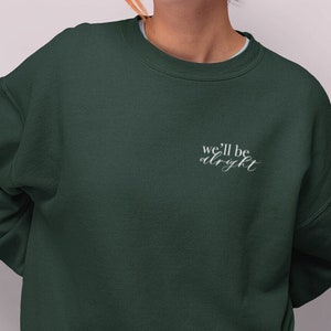 Peut inclure: Sweat-shirt vert foncé avec l'inscription brodée "we'll be alright" en écriture cursive. Le sweat-shirt est de couleur unie et présente une encolure ras du cou.