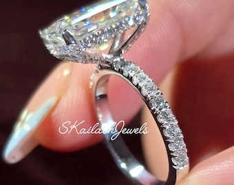 May include: A diamond ring with a large, rectangular-cut central stone, surrounded by a halo of smaller diamonds. The band is also set with diamonds. The ring is held by a person. The ring is silver in colour.